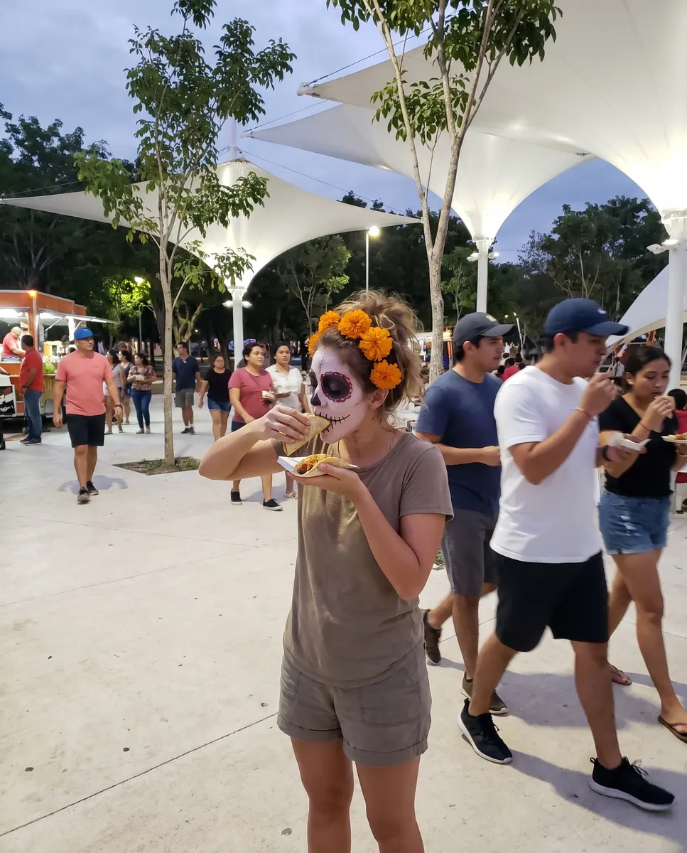 Mujer con maquillaje de catrina y cempasúchil en el pelo, comiendo un taco entre la gente